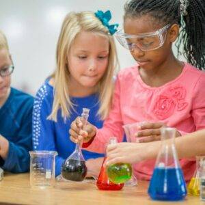 A group of children performing science experiments. Two are wearing safety goggles and there are beakers of coloured liquid in front of them.