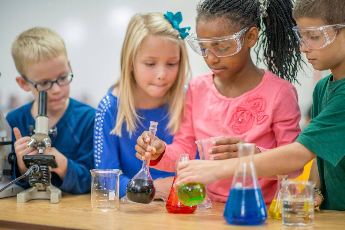 A group of children performing science experiments. Two are wearing safety goggles and there are beakers of coloured liquid in front of them.