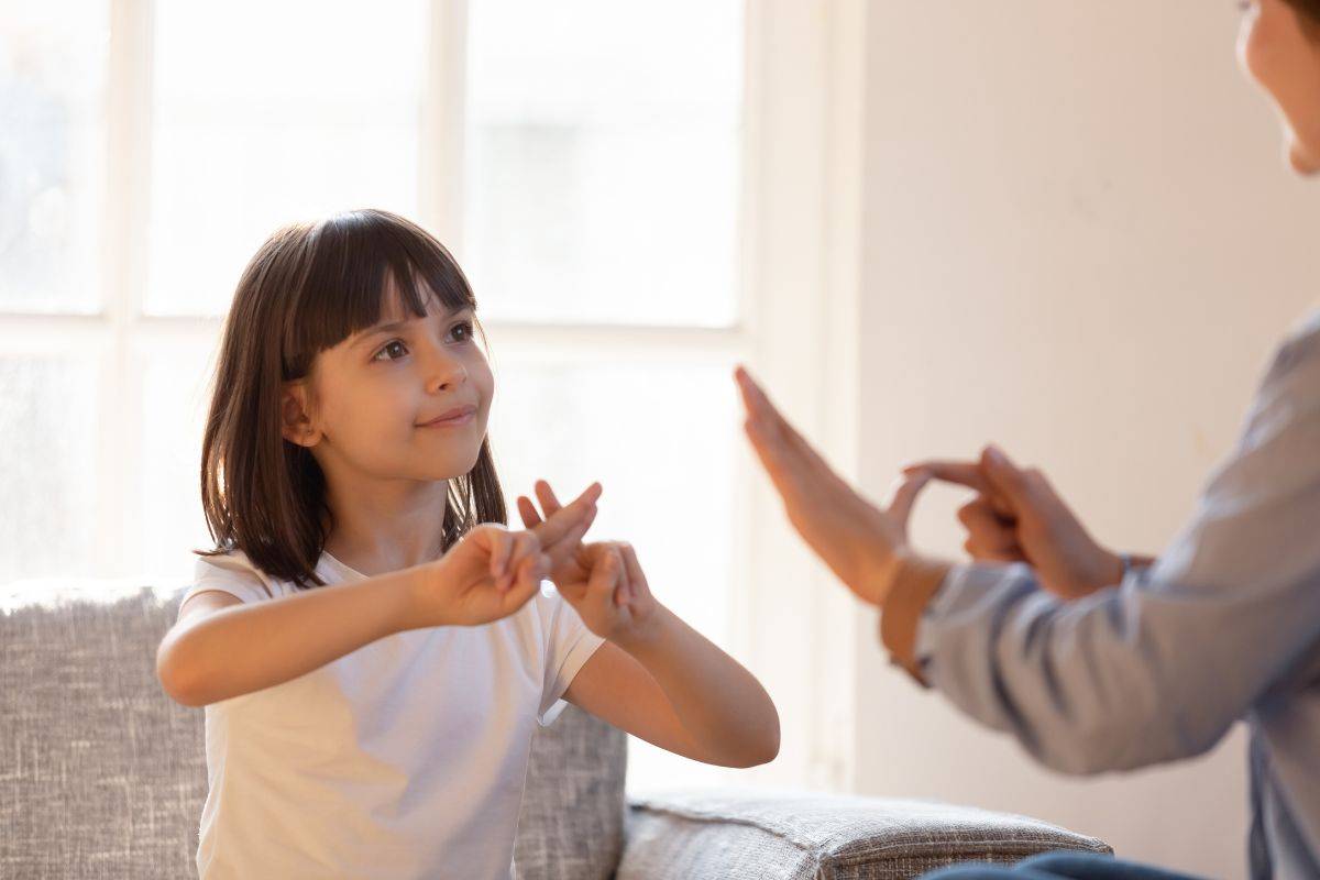 A young girl is sitting down on a sofa, using sign language to communicate with an adult.