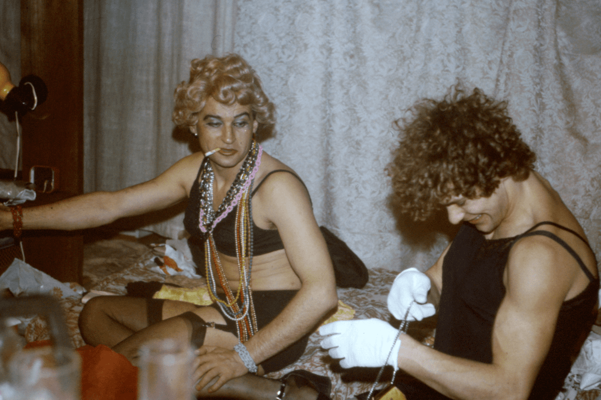 Two people dressed in black lingerie and putting on costume jewellery while smiling.