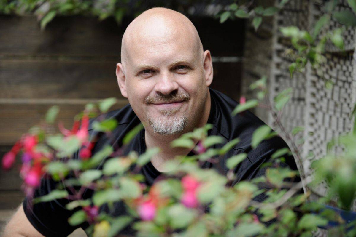 A man seated outside in a garden, in front of him is a small bush. He is smiling.