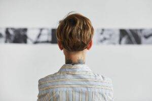 The back of someone's head looking at a wall of art at an exhibition. They have short brown hair and are wearing a white shirt. They have a tattoo running up the back of their neck.
