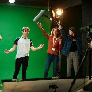 A television studio with a group of people doing various jobs. There is a presenter, a cameraperson, a light technician and someone with a clapper board. Behind them is a green screen.