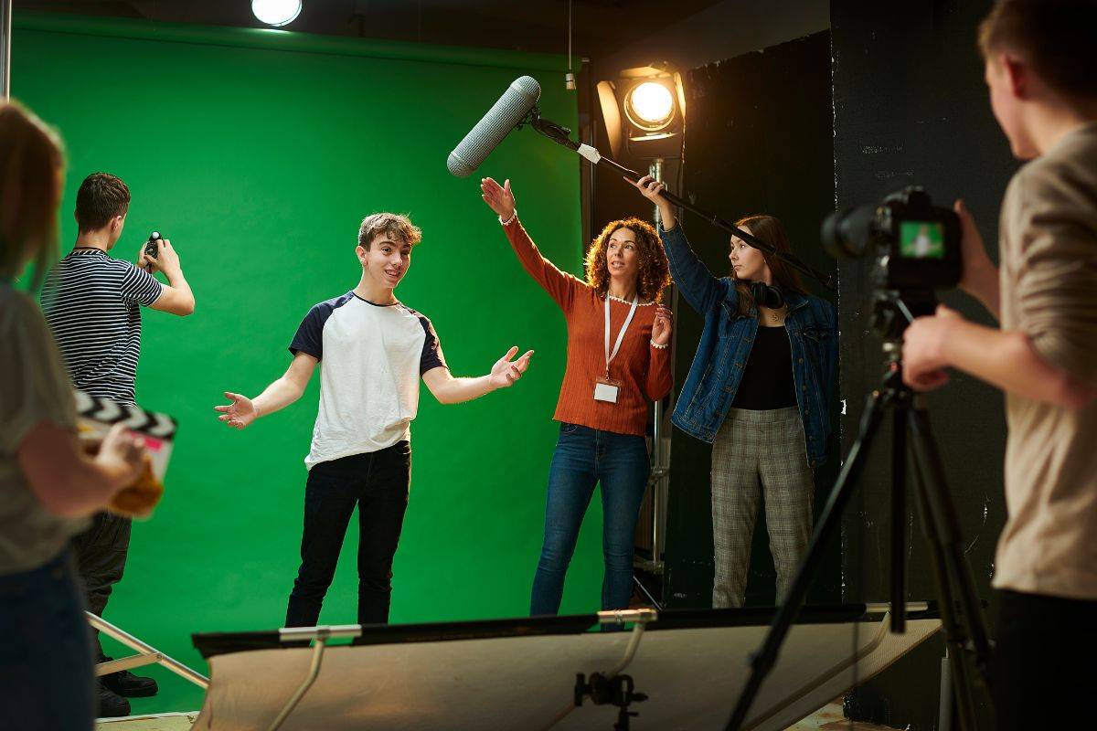 A television studio with a group of people doing various jobs. There is a presenter, a cameraperson, a light technician and someone with a clapper board. Behind them is a green screen.