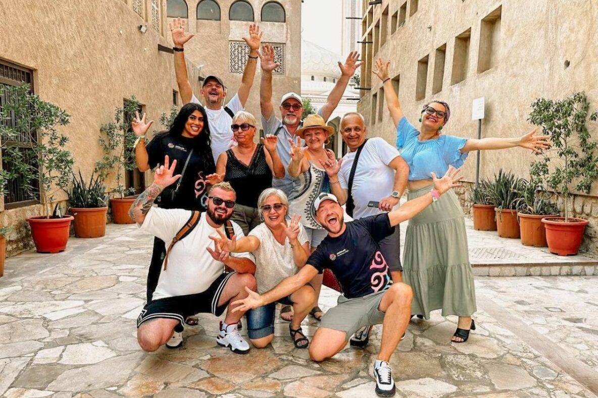 A group of people posing for a photo. Three are kneeling down and the front and the rest are behind them with their hands in the airs and smiling. There are on a street with sand coloured buildings and a stone floor.