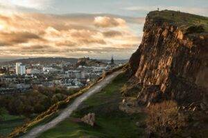 An image of Holyrood park in Edinburgh at dusk. There is a large mountain in the foreground and Edinburgh city in the background.