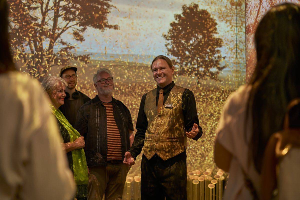 A group of people stand around looking at a man dressed in a golden waistcoat as he is giving a tour. Behind him is an image of a barley field.