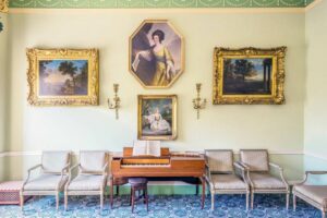 A georgian room with paintings on the wall, a desk and four chairs.