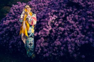 Linder standing in front of a purple bush holding a yellow scarf.