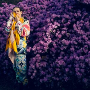 Linder standing in front of a purple bush holding a yellow scarf.