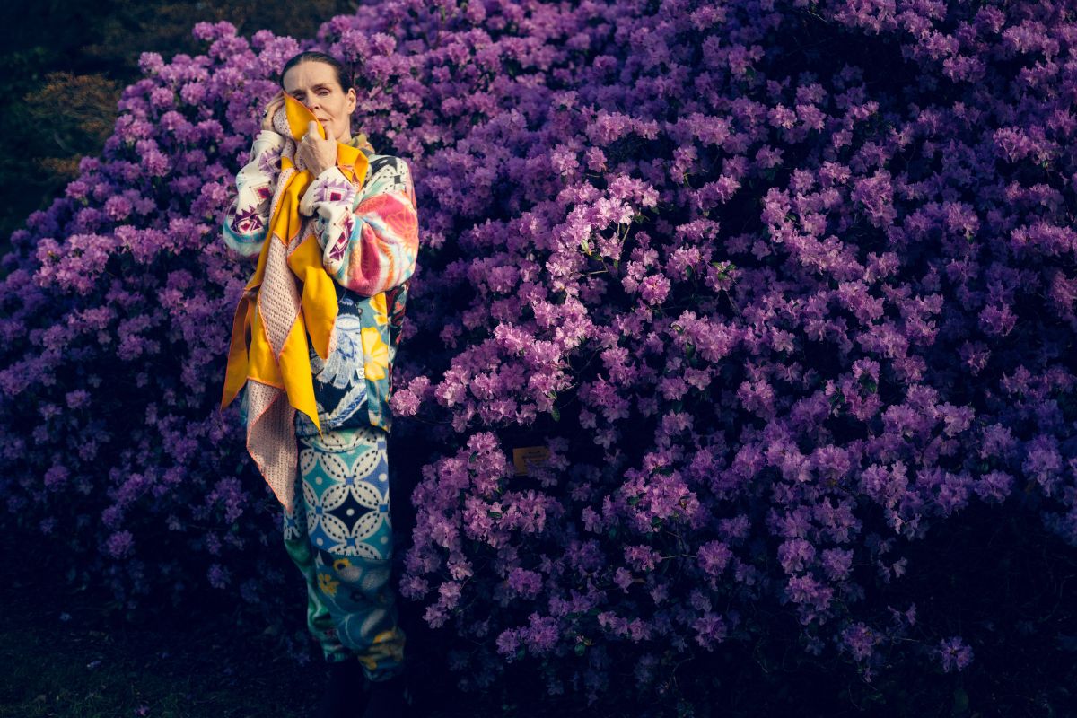 Linder standing in front of a purple bush holding a yellow scarf.