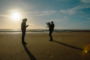 Two people standing on a beach, one his holding a camera and pointing it at the other.