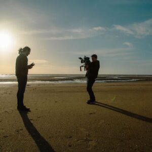 Two people standing on a beach, one his holding a camera and pointing it at the other.