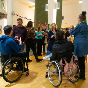 A group of people standing in a room networking. There are two people in wheelchairs and other people standing using sign language to communicate.