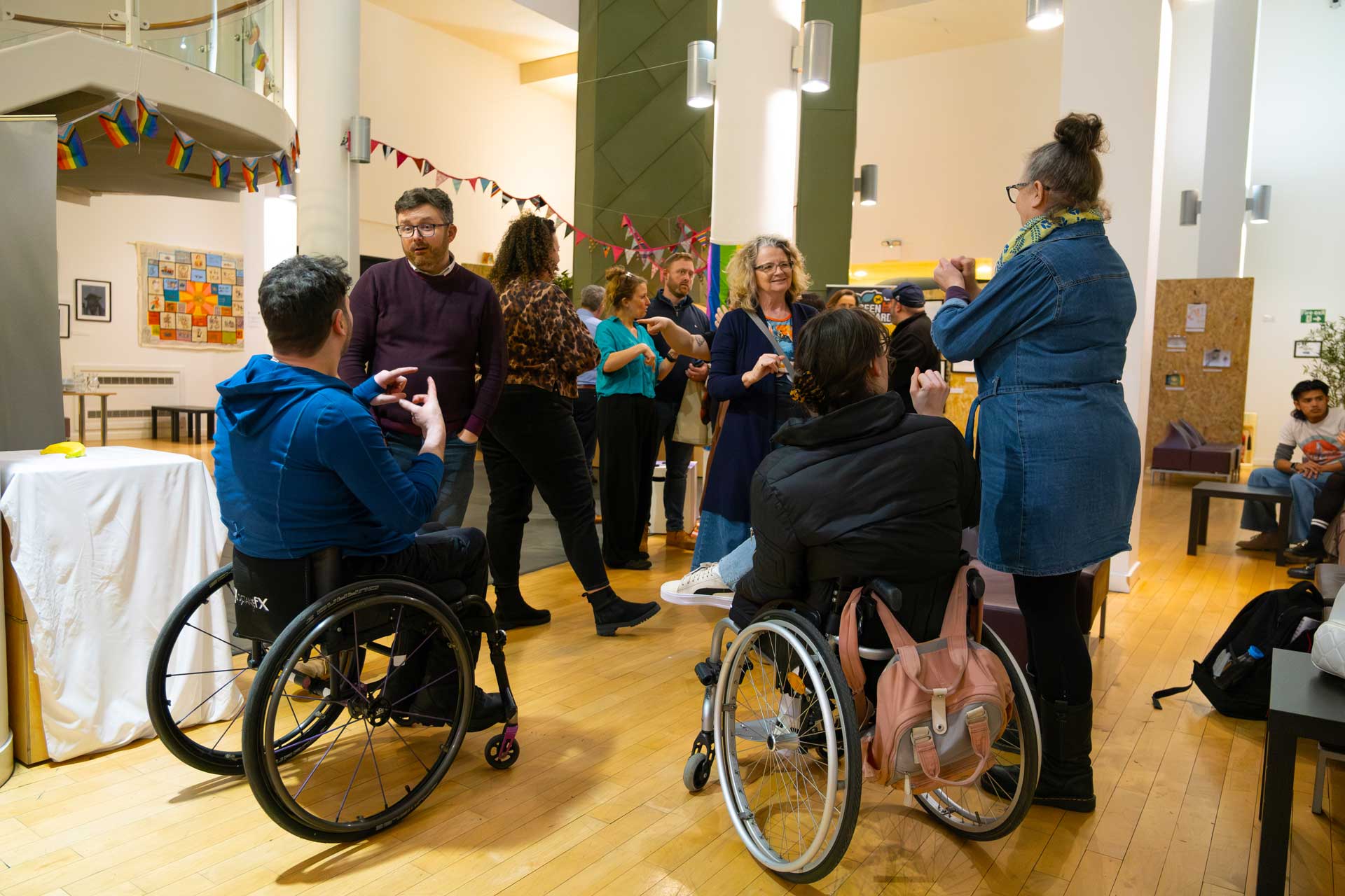 A group of people standing in a room networking. There are two people in wheelchairs and other people standing using sign language to communicate.