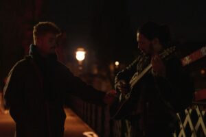 Two men standing beside each other, one is holding a guitar.