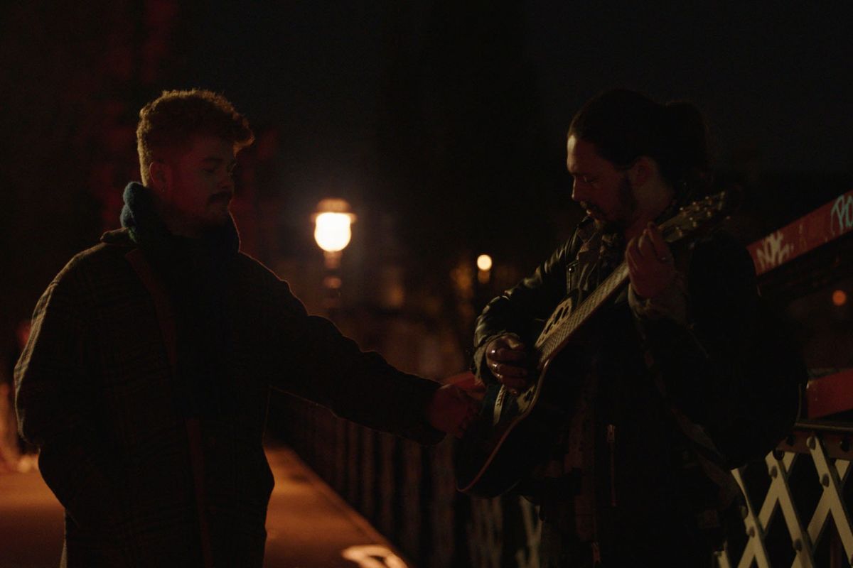 Two men standing beside each other, one is holding a guitar.